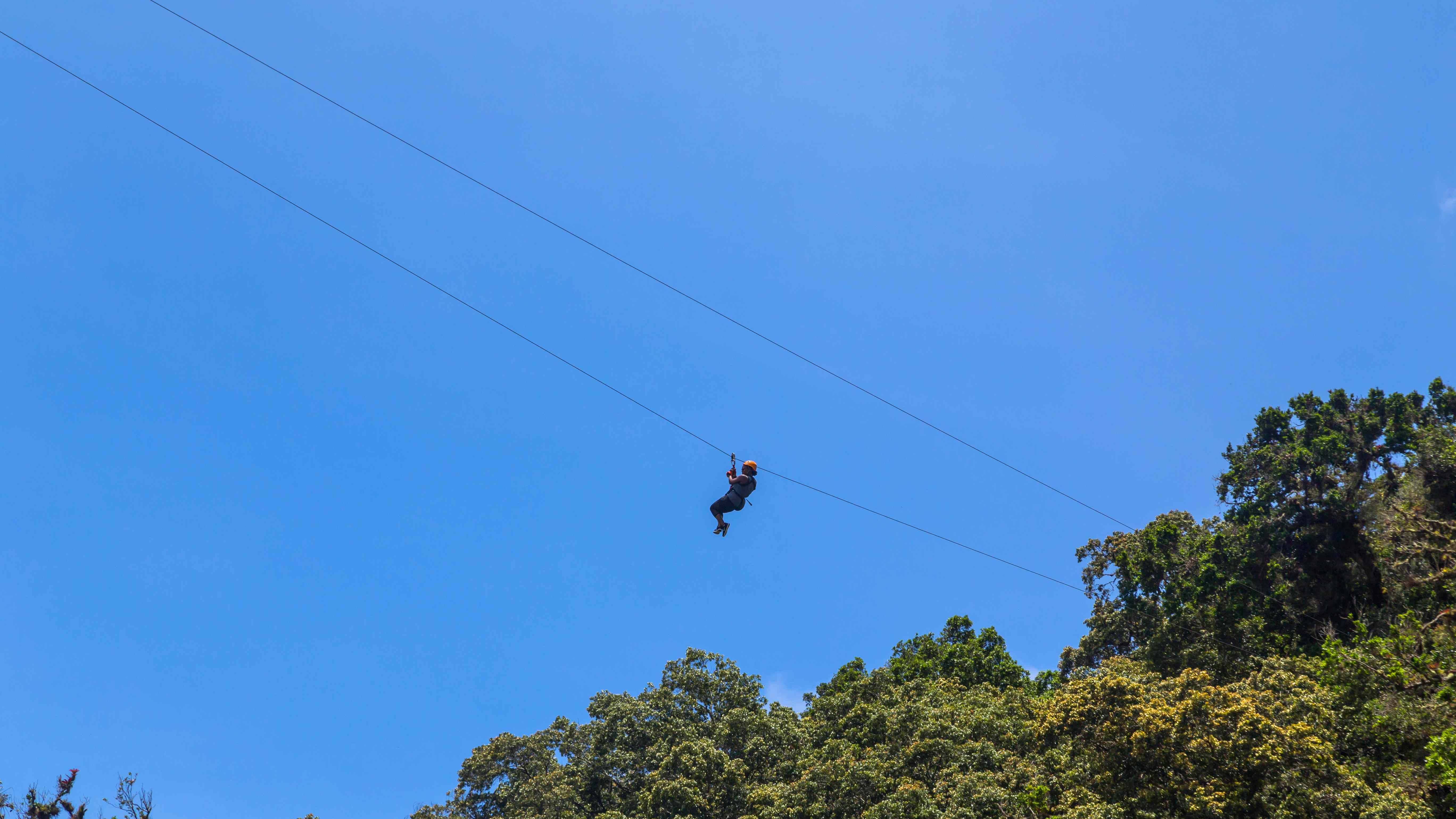 Persoon aan het ziplinen boven het nevelwoud van Monteverde, met een heldere blauwe lucht op de achtergrond.